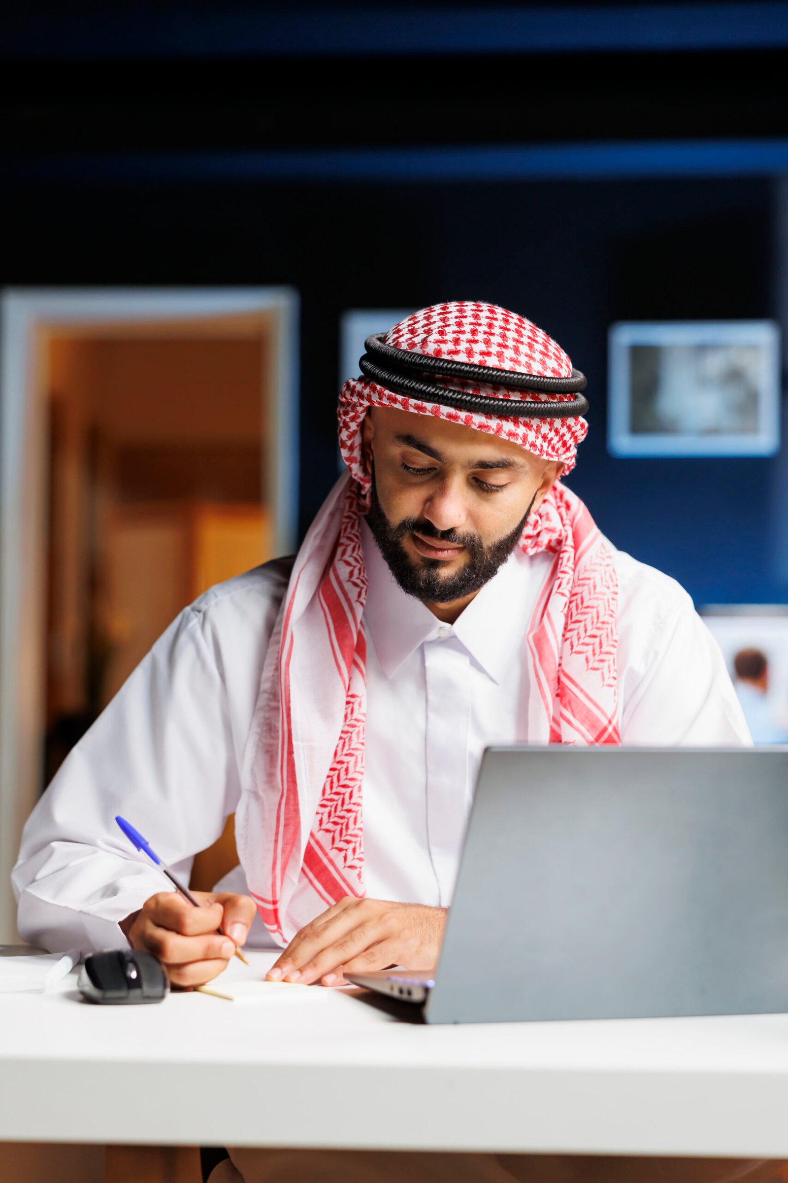 An Arab businessman, absorbed in work, diligently browses digital information on office desk laptop. He takes notes while conducting internet research, demonstrating his professionalism and attention.